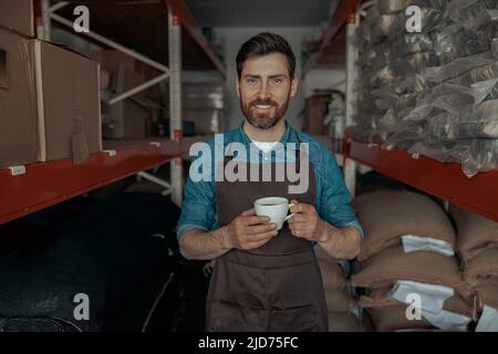 Smiling barista in uniform drinking coffee on warehouse with bags of coffee beans  Stock Photo