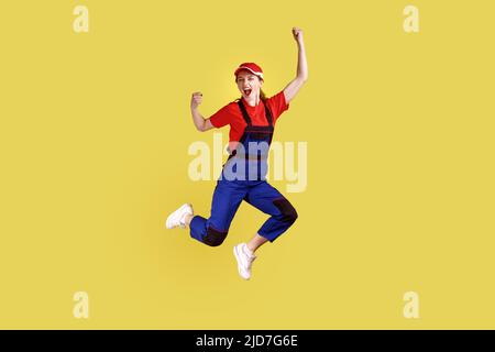 Full length portrait of extremely happy worker woman jumping and clenched fists, celebrating long awaited day off, wearing overalls and red cap. Indoor studio shot isolated on yellow background. Stock Photo