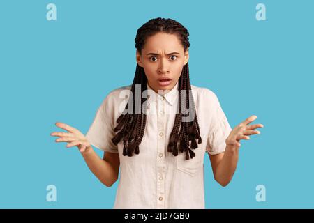 How could you. Portrait of annoyed frustrated woman with black dreadlocks standing with raised hands and indignant face asking why, wearing white shirt. Indoor studio shot isolated on blue background. Stock Photo