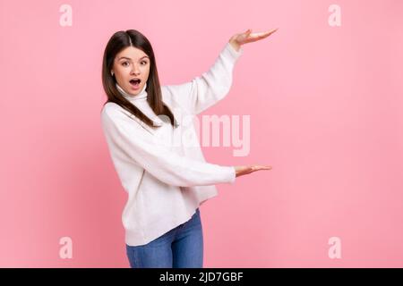 Woman presents copy space between hands, looks surprised, holds empty place for commercial idea, wearing white casual style sweater. Indoor studio shot isolated on pink background. Stock Photo