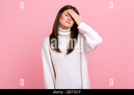 Unlucky girl standing with facepalm gesture, feeling regret and sorrow, blaming herself for mistake, wearing white casual style sweater. Indoor studio shot isolated on pink background. Stock Photo