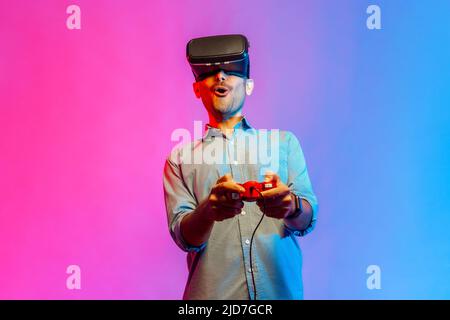 Portrait of amazed man in shirt in VR glasses playing video game, using simulator, yelling emotionally, being excited of gaming. Indoor studio shot isolated on colorful neon light background. Stock Photo