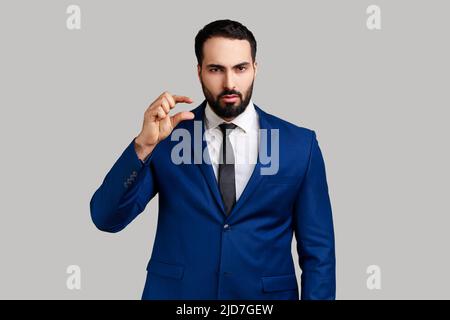 Bearded man looking disappointed and showing a little bit gesture, small amount or low scale sign, few centimeters, wearing official style suit. Indoor studio shot isolated on gray background. Stock Photo