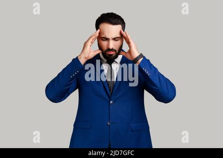 Bearded man frowning and clasping sore head, suffering intense headache, having unbearable migraine, fever and flu symptoms, wearing official style suit. Indoor studio shot isolated on gray background Stock Photo