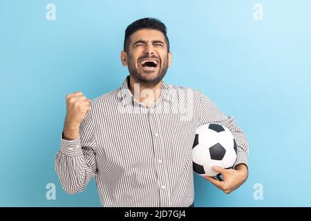 Excited businessman screaming widely opening mouth, celebrating victory of favourite football team, holding soccer ball in hands, wearing striped shirt. Indoor studio shot isolated on blue background. Stock Photo