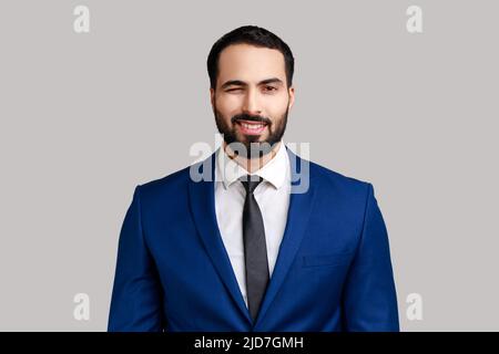 Portrait of bearded businessman in good mood, smiling broadly and winking at camera with toothy smile, wearing official style suit. Indoor studio shot isolated on gray background. Stock Photo