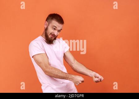 Portrait of strong bearded man keeping fists up, holding invisible virtual rope and pulling object, striving hard with effort, wearing pink T-shirt. Indoor studio shot isolated on orange background. Stock Photo
