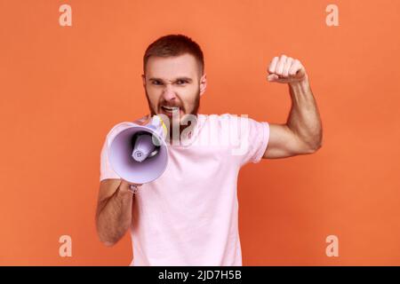 Portrait of angry bearded man loudly screaming at megaphone, making announce, protesting, wants to be heard, wearing pink T-shirt. Indoor studio shot isolated on orange background. Stock Photo