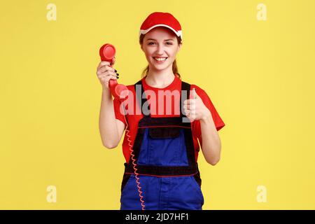 Portrait of delighted worker woman holding out handset, showing thumb up, likes order service, looking friendly at camera, wearing overalls and cap. Indoor studio shot isolated on yellow background. Stock Photo