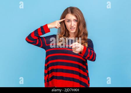 Hey, you are kidding. Portrait of young adult beautiful woman wearing sweater, holding finger near head and pointing finger at camera, stupid gesture. Indoor studio shot isolated on blue background. Stock Photo