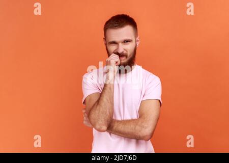 Portrait of stressed out, worried bearded man biting nails, nervous about troubles, panicking and looking scared, wearing pink T-shirt. Indoor studio shot isolated on orange background. Stock Photo