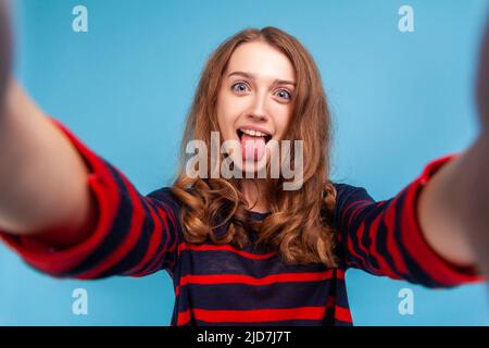 Childish woman wearing striped casual style sweater, having funny facial expression taking selfie and showing tongue out, point of view of photo.Indoor studio shot isolated on blue background. Stock Photo