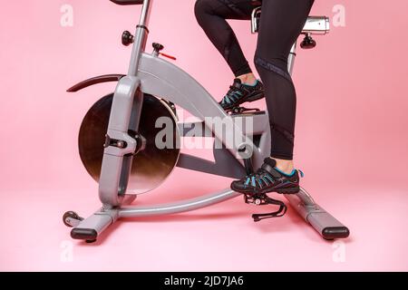Portrait of the bike simulator and women's legs in black sneakers and leggins, cardio workout, training. Indoor studio shot isolated on pink background. Stock Photo