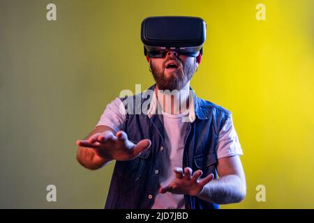 Amazed hipster man standing in virtual reality headset, playing video game and trying to touch something, wearing hat and denim vest. Indoor studio shot isolated on colorful neon light background. Stock Photo