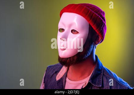 Side view of anonymous hipster man in white mask on face, hiding his identity, spying undercover, corruption, wearing hat and denim vest. Indoor studio shot isolated on colorful neon light background. Stock Photo