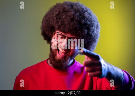 Hipster man with Afro hairstyle pointing finger on you and winking at camera, you are special, victory in lottery, chance of success. Indoor studio shot isolated on colorful neon light background. Stock Photo