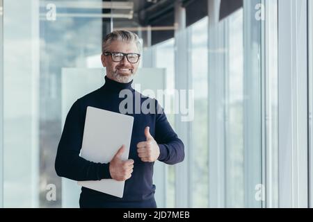 Portrait of senior and experienced teacher, gray-haired university teacher by the window smiling and looking at camera holding laptop in hand and show Stock Photo
