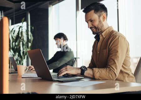 Happy male company employee using laptop, checking email, typing on keyboard, working with male colleague at office Stock Photo