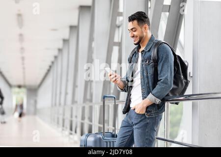 Smiling Arab Male Traveller With Smartphone And Luggage Waiting In Airport Stock Photo