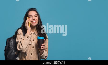 Smiling millennial caucasian female brunette in casual with backpack hold coffee cup takeaway and talking on phone Stock Photo