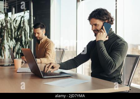 Joyful young businessman using laptop, talking on cellphone, working with male colleague at company office, free space Stock Photo