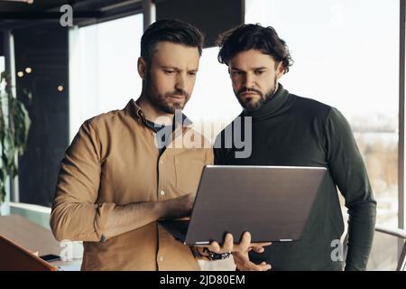 Two serious male employees developing business strategies, coworking together, using laptop at contemporary office Stock Photo