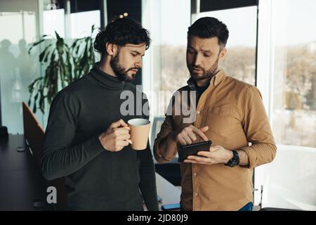 Portrait of two serious young colleagues using digital tablet, discussing business project, drinking coffee at office Stock Photo