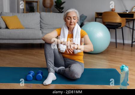 Senior woman using smartwatch fitness tracker after domestic workout, checking burned calories, sitting on yoga mat Stock Photo