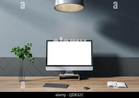 Front view on computer monitor with white blank screen on dark wooden table with black keyboard, notebook and glass vase in sunny room with dark wall. Stock Photo