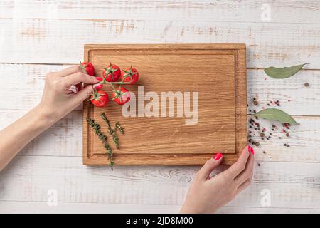 hands of a woman holding a square wooden cutting board with cherry tomatoes and spices on a white background. mockup with copy space for text, top vie Stock Photo