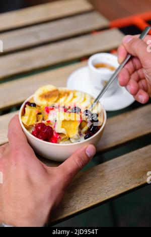 Close up vertical photo male hands holding a bowl on top of wooden table, scooping waffle bowl full with milk, raspberry, blueberries, mini waffles an Stock Photo