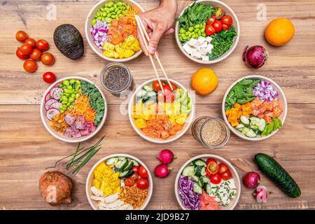 Set of large white bowls filled with salmon, chicken, bluefin tuna, lots of mango, cherry tomatoes, cucumbers, sweet potato, red onion and a hand with Stock Photo