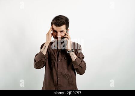 Portrait of young handsome man wearing brown shirt posing isolated over white background feeling upset desperate talking on the phone having problems. Stock Photo