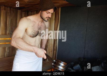 Brutal man in sauna pouring water from ladle on hot stones Stock Photo