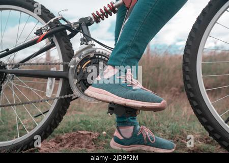 Detail of the feet of a young woman wearing blue pants and blue and pink sports shoes on the pedals of her bicycle in the middle of the grassy field o Stock Photo