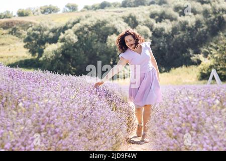 Soft selective focus girl in purple dress walks on lavender field and enjoys the aroma of pink flowers and beauty on sunny day. Trendy color of 2022 Very peri. Digital detox pleasure of slow life. Stock Photo