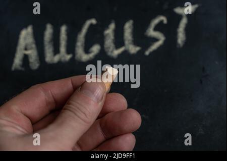 Adult male holding a piece of yellow chalk in his hand. Handwritten word AUGUST on black chalkboard. Selective focus. Stock Photo