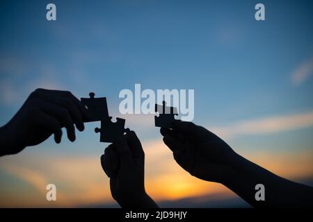 Close-up silhouette of hands holding and putting pieces of a white puzzle together against the background of the sunset sky. The concept of team Stock Photo