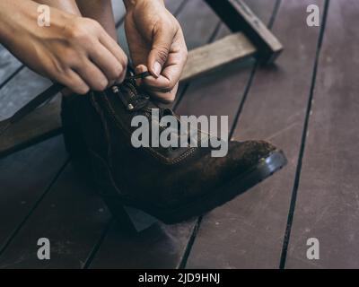 Close-up woman's hands tying shoelace on the old leather boots in her legs while sit on wooden chair on the wood plank terrace. Stock Photo