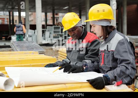 Two confident builders in hardhats and workwear discussing sketch on blueprint at working meeting while one of them pointing at it Stock Photo