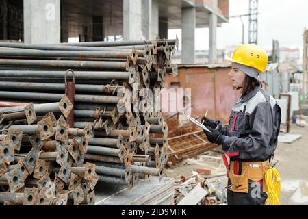 Young female engineer in workwear making notes in document while standing in front of two stacks of metallic pipes at construction area Stock Photo
