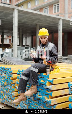 Young female builder in workwear having sandwich and using smartphone while sitting on stack of planks at lunch break Stock Photo