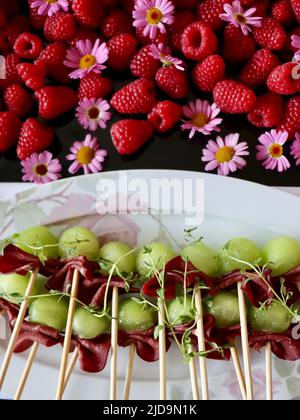 Bamboo skewers with smoked reindeer and melon on tray decorated with raspberries and purple flowers. Stock Photo