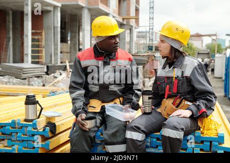 Two modern engineers or builders with cups of hot tea having talk at construction site while sitting on stack of yellow building materials Stock Photo
