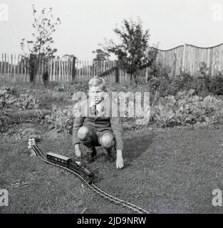 1940s, historical, a young schoolboy kneeling down outside on a lawn for a photo next to his wind-up train on a track on the grass, Bramhall, Stockport, England, UK. Stock Photo