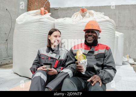 Cheerful intercultural workers of construction site having lunch against sacks with concrete or other building materials and chatting at break Stock Photo