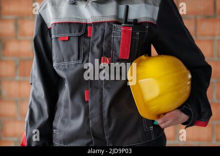 Middle section of young modern female builder in uniform holding protective helmet of yellow color while standing in front of camera Stock Photo
