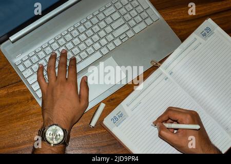 Top view of businessman typing on laptop while writing on a diary on workplace. Selective focus on laptop keyboard. Stock Photo
