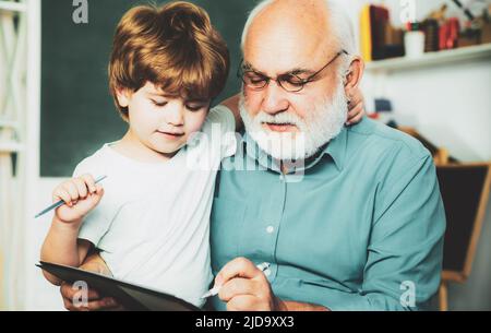 Grandfather with grandson learning together. Friendly child boy with old mature teacher in classroom near blackboard desk. Teacher is skilled leader. Stock Photo
