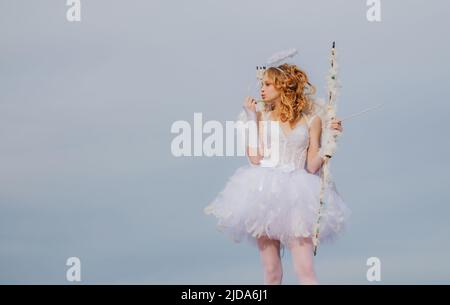 Angel teen with arrow and wings. Charming curly little girl in white dress and wings - angel cupid girl. St Valentines day. Space for text. Postcard Stock Photo